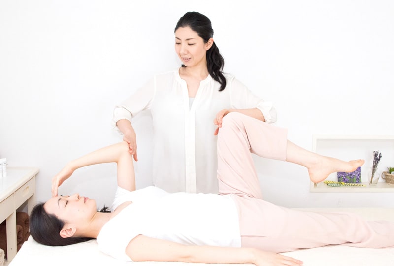 仰向けに横たわる女性の肘と膝に触れるIMSI副学院長　嵯峨 慈子