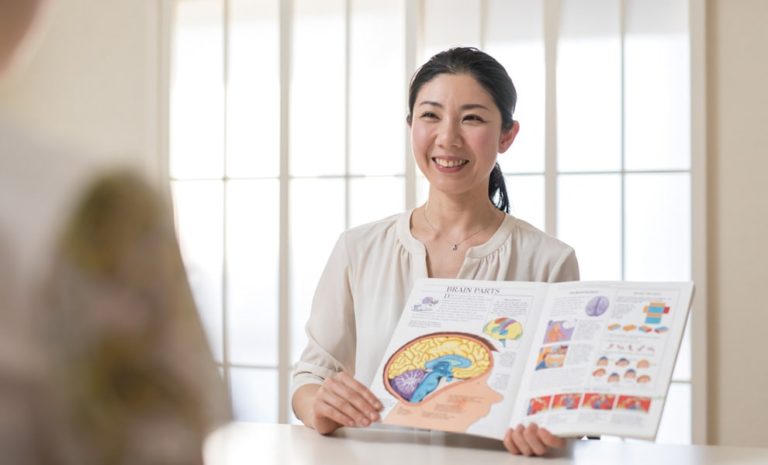 脳に関する本を広げながら説明するIMSI副学院長　嵯峨 慈子